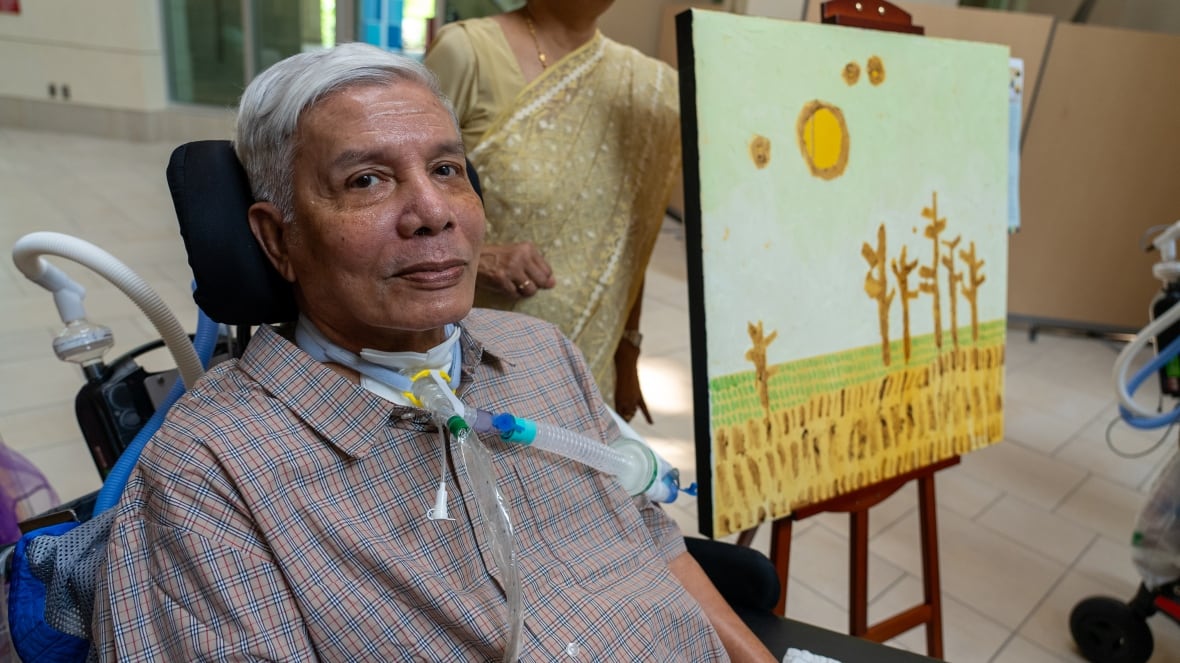 Mr. Kazi, a long-term care patient, sits beside his artwork—a peaceful scene of trees, sunlight, and grassy fields.