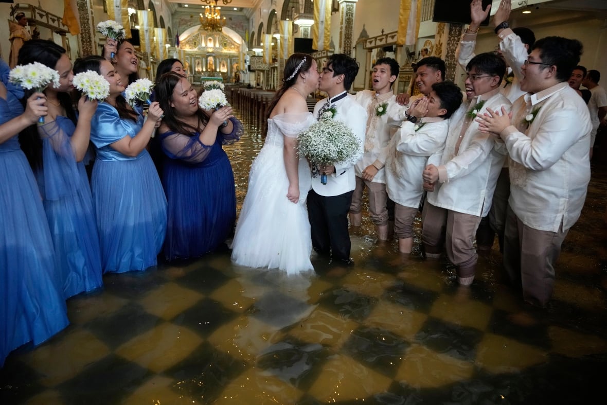 A man and woman kiss while surrounded by women in blue dresses and men in suits while water covers the floor.