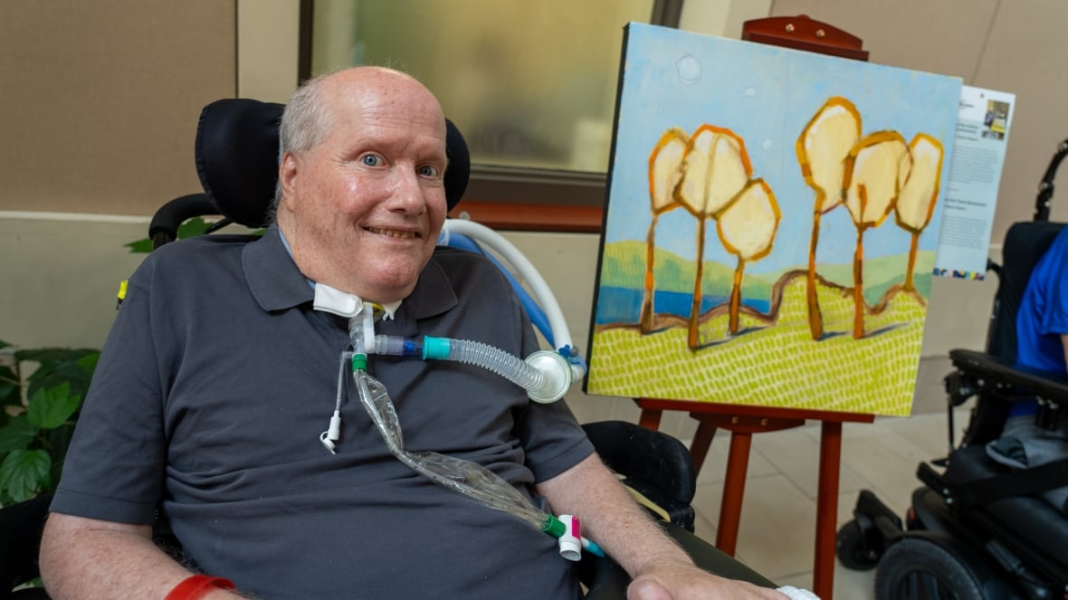Patient Martin Meech sits beside his painting Where the Trees Remember, which shows six trees on grassy land near a lake and rolling hills.