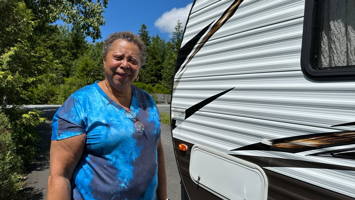 A woman wearing a blue t shirt smiles at the camera standing by a camping trailer she is taking to the reunion in Africville.