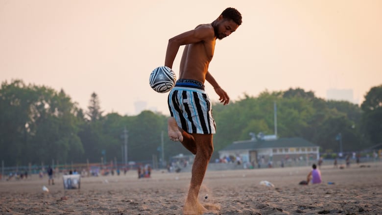 A man in striped shorts kicks a soccer ball on a hazy beach at sunset, with other beachgoers seated in the background.