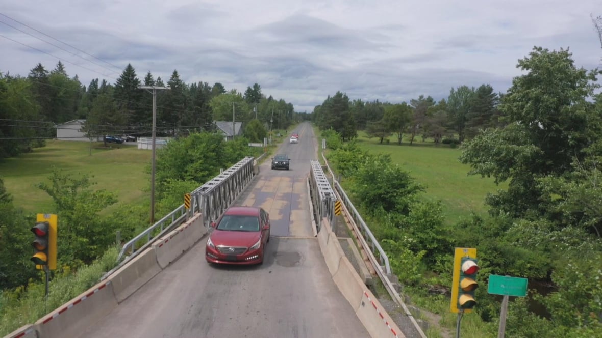 An overhead view of cars crossing a narrow replacement bridge over a shallow river.