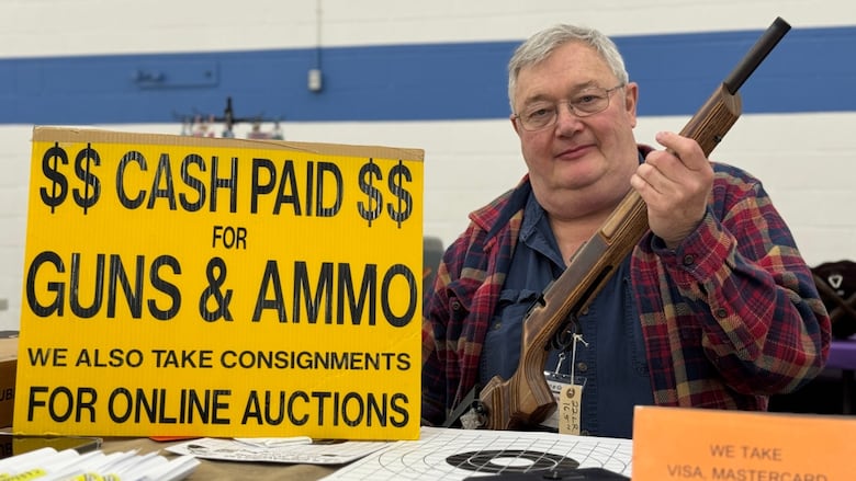 A man holds up a gun in front of a sign that reads, Cash paid for guns and ammo.