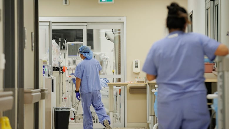 Two nurses wearing blue scrubs stand in a hallway across from each other. 