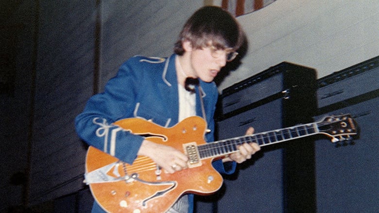 A person in a blue jacket plays an orange electric guitar. 