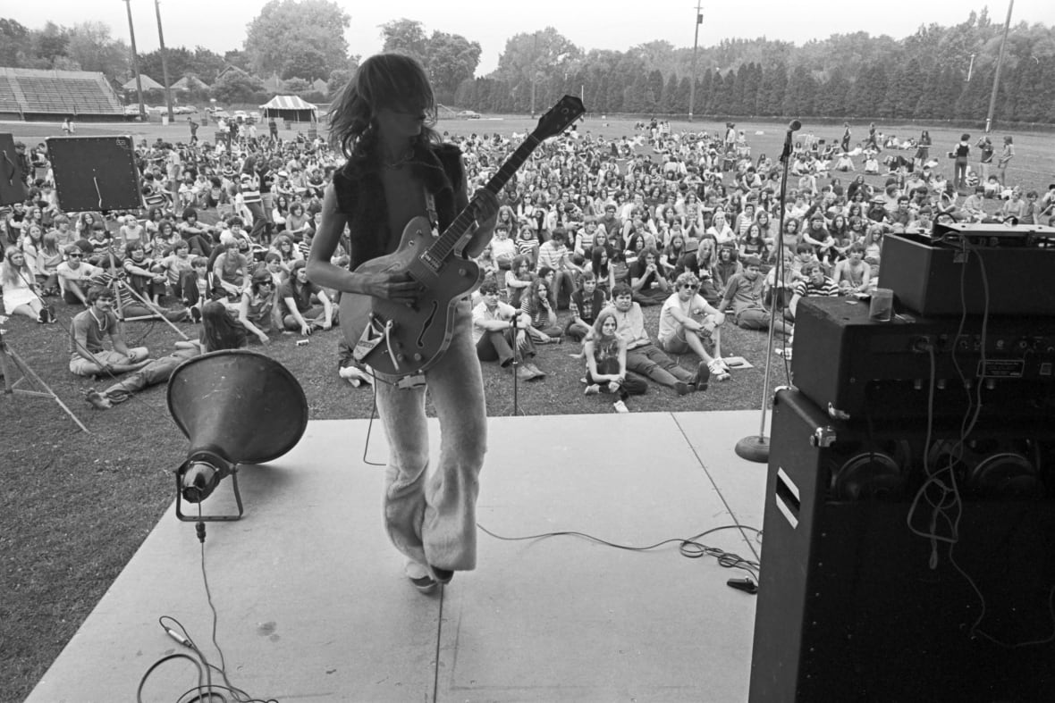 A black and white image of a person with long hair playing guitar on stage facing the camera with their back to an audience on a lawn outside.