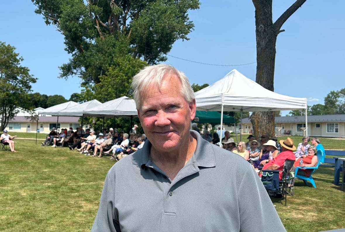 Man wears grey golf shirt as people sit under tents in background.