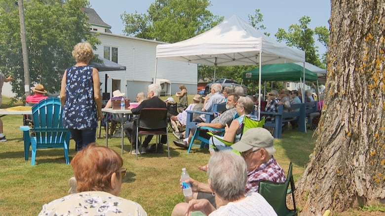 People sit in chairs and under tents outside on a sunny day.