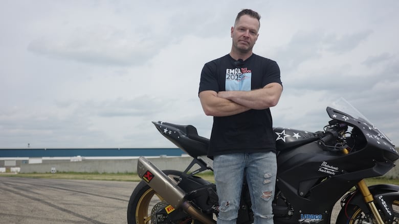 Jon Bullee stands in front of his black Honda racing motorcycle with a track littered with burnt rubber marks behind him. 
