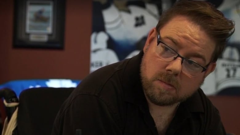 A man with short brown hair and glasses, wearing a short-sleeved black shirt, sits in a wheelchair and looks to the side.