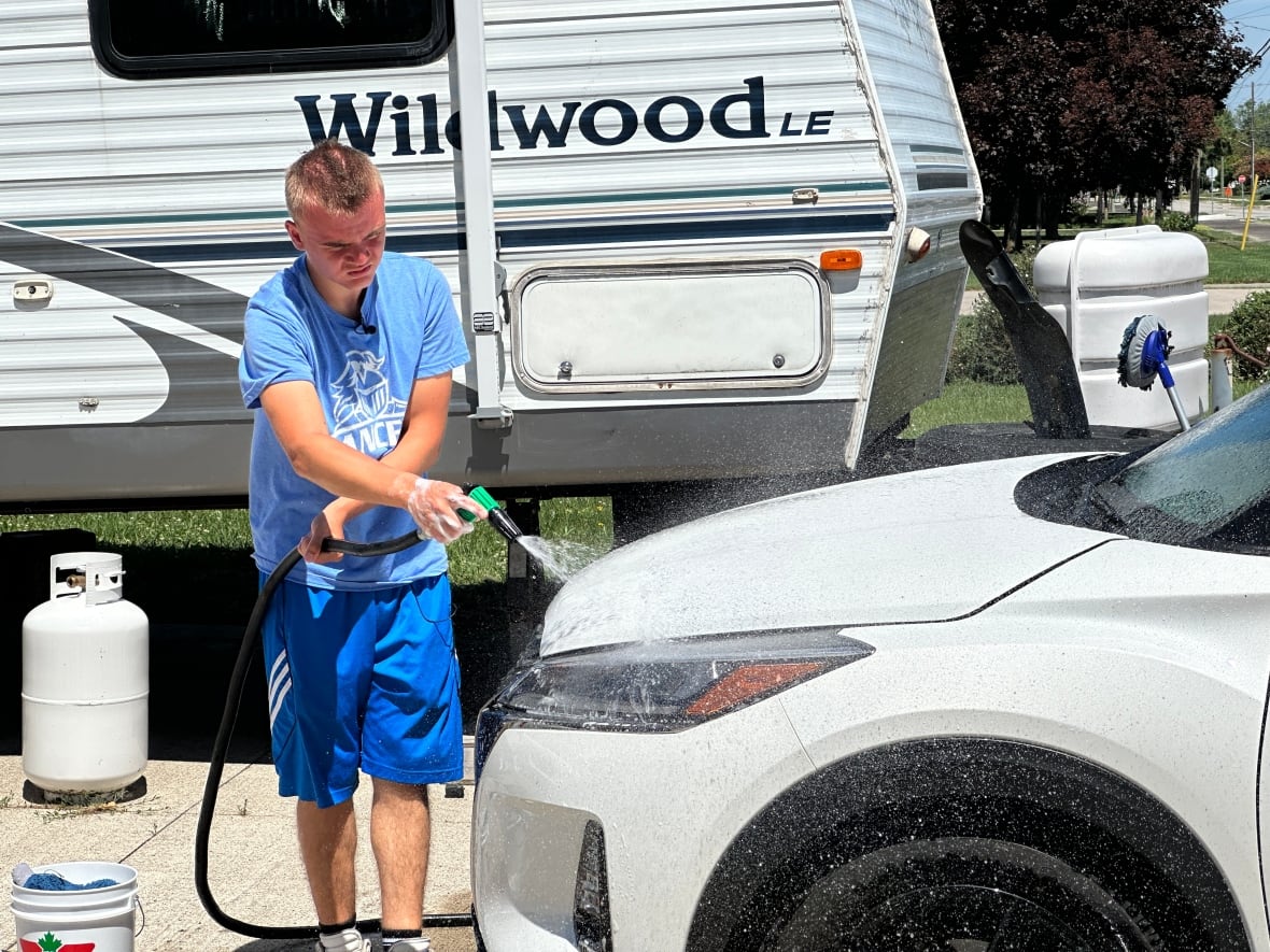 A slim teenager in a t-shirt and jean shorts sprays the front of an SUF with a hose.  