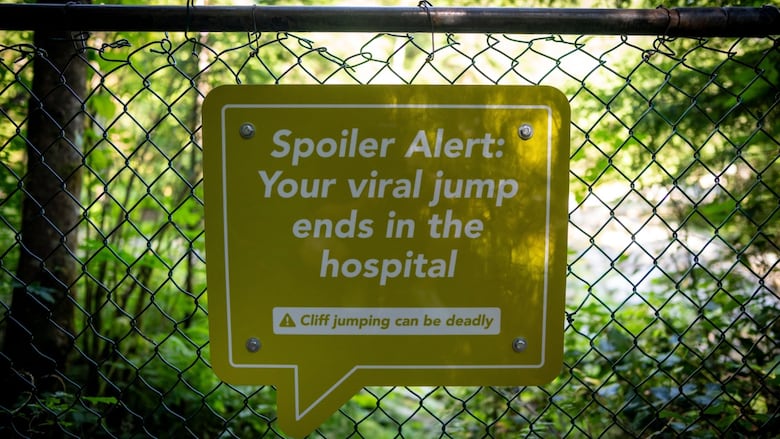 A yellow-green sign attached to a park fence reads, 'Spoiler Alert: Your viral jump ends in the hospital.'