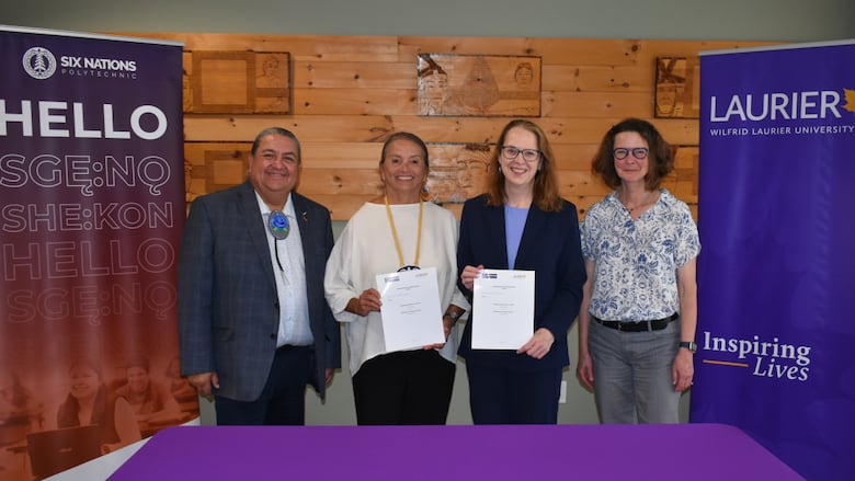 People standing and holding documents while looking at the camera. A banner for Six Nations Polytechnic is on one side and a banner for Wilfrid Laurier University is on the other. 
