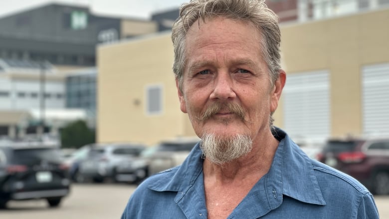 A grey-haired man in a blue shirt stands outside of a hospital building.