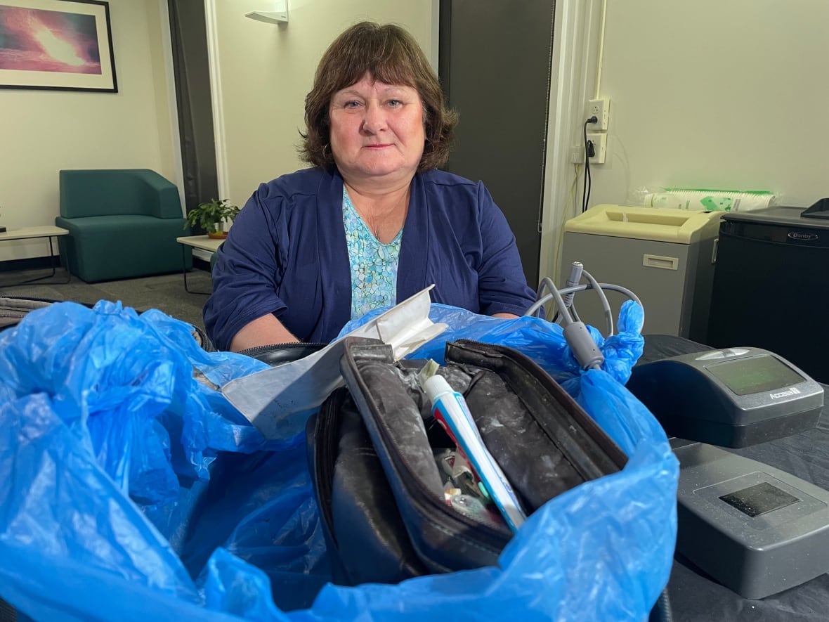 Woman seated at a table, with a suitcase laid in front of her. Inside the open bad is a black toiletry bag and a scanning device.
