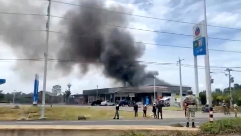 Large black smoke is shown from a distance rising above a gas station. Dozens of people are shown standing and staring at the smoke.