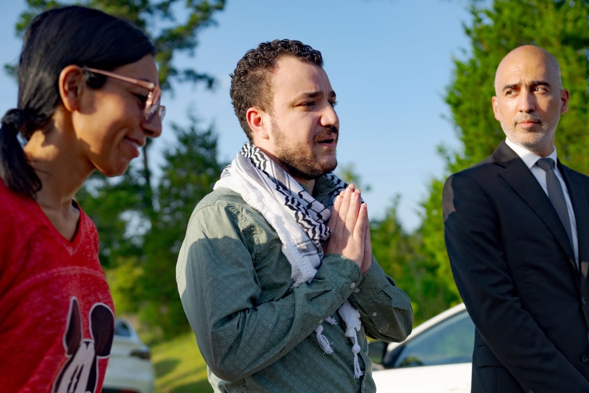 A bearded dark haired man is shown speaking, he is flanked by a woman in glasses to his right, and an older man to his left.