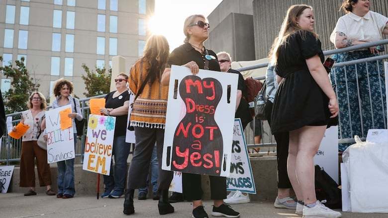 A group of people stand with signs on a sidewalk on a sunny day.