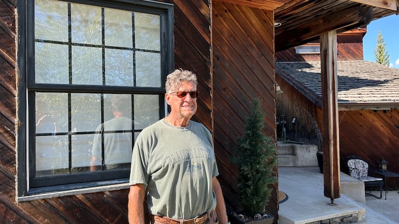 A man in a green T-shirt and brown shorts stands in front of a wood house.