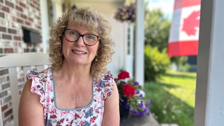A woman with curly blonde hair, glasses and a floral sun dress smiles at the camera while sitting on a front porch on a summer day.