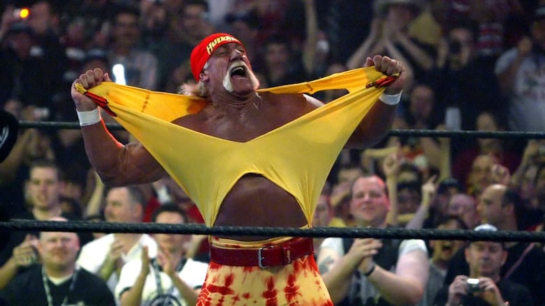 A man in a bandana, heavily tanned and with a mustache, rips his shirt inside a wrestling ring.
