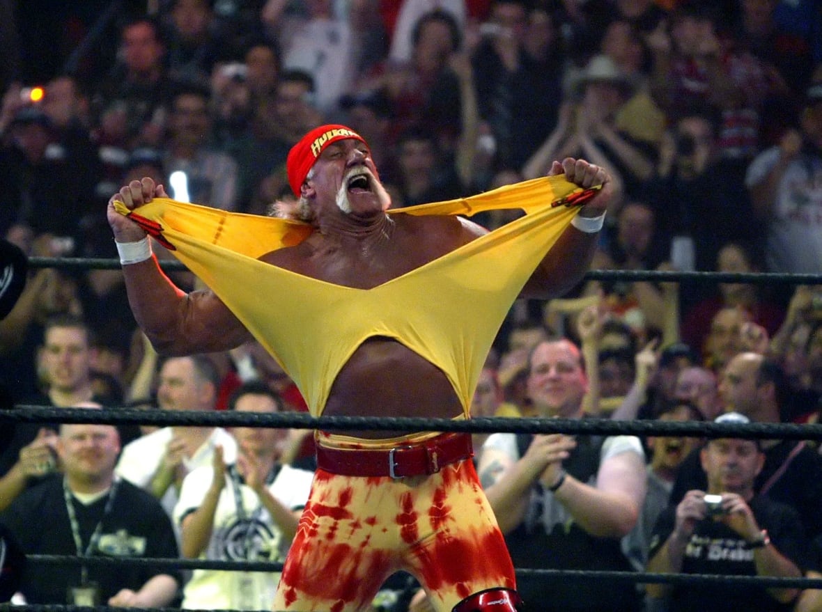A man in a bandana, heavily tanned and with a mustache, rips his shirt inside a wrestling ring.