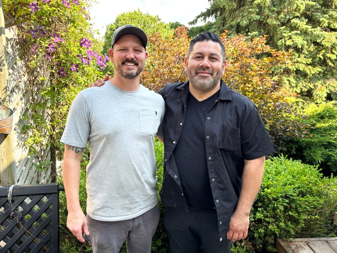 Two men stand side by side. One is wearing a grey shirt, the other is in black.
