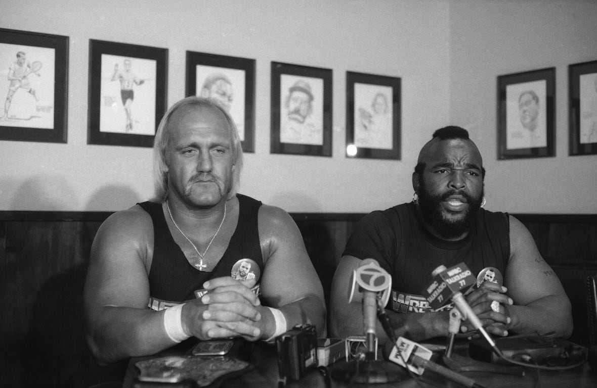 In a black and white photo, two men sit in a booth with recording microphones set up on the table.