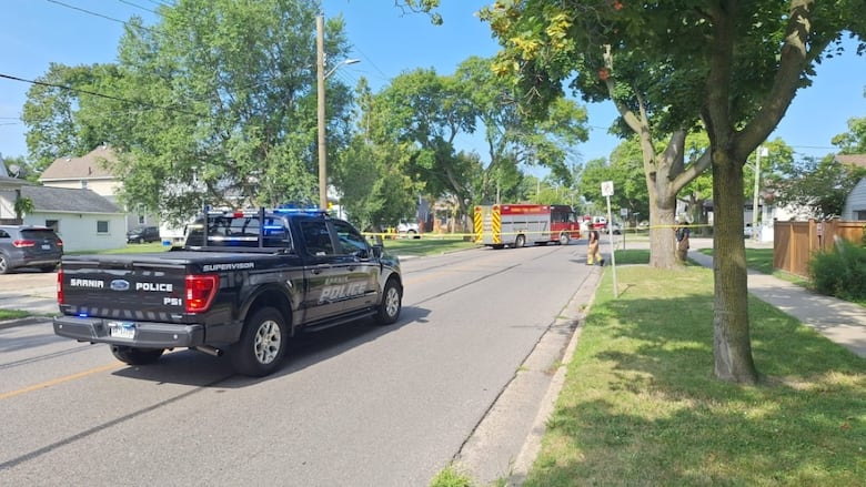 A Sarnia police truck in the foreground parked in the middle of the road.  A fire truck seen farther down the road.  A line of yellow tape runs across the road between them.