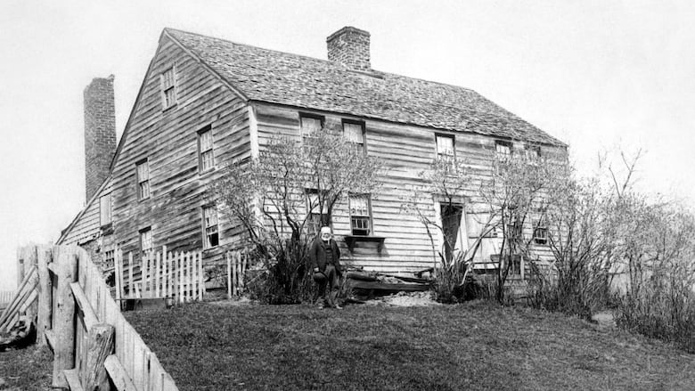 An old black and white photo of a two-storey home with a man standing in front of it. 