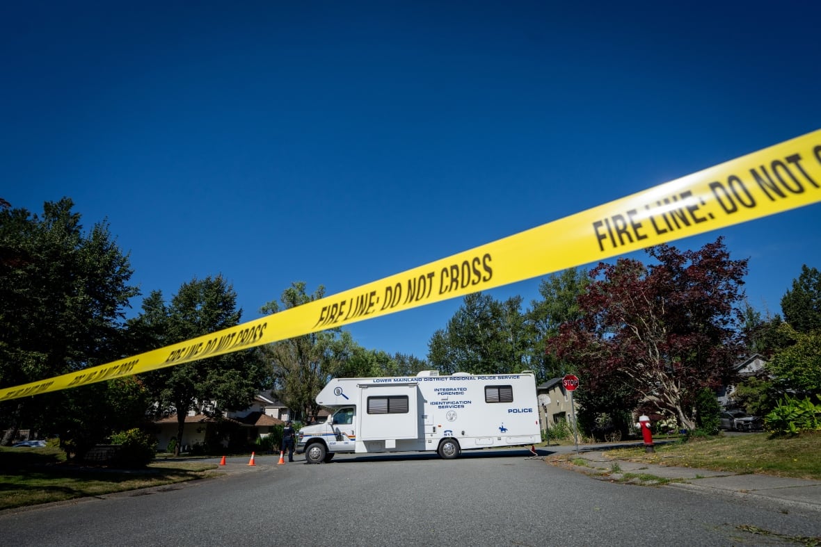 Yellow police tape stretches across a road with a large white vehicle parked in the middle.