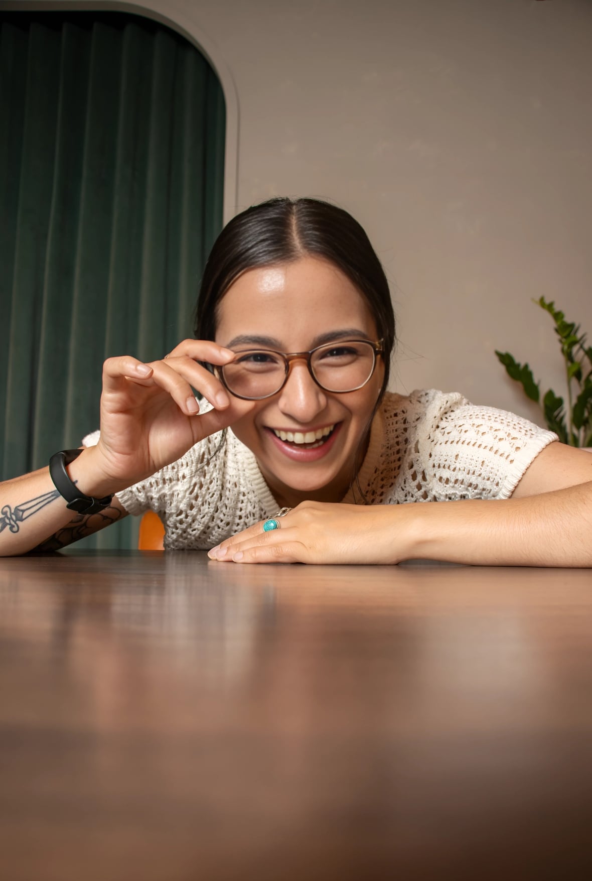 A woman with dark hair adjusts her glasses with her arms resting on a table.