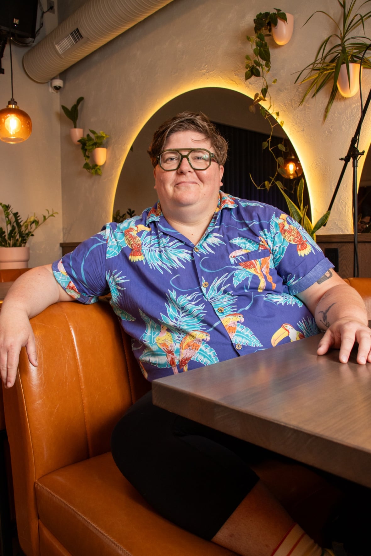 A person with medium-length hair, glasses and a printed shirt sits at a booth.