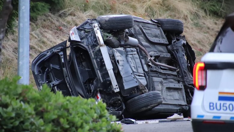 A vehicle that appears to be a black SUV is turned over on its side, its underside visible in the image. The corner of a white police vehicle is seen on the bottom right-hand corner of the image.