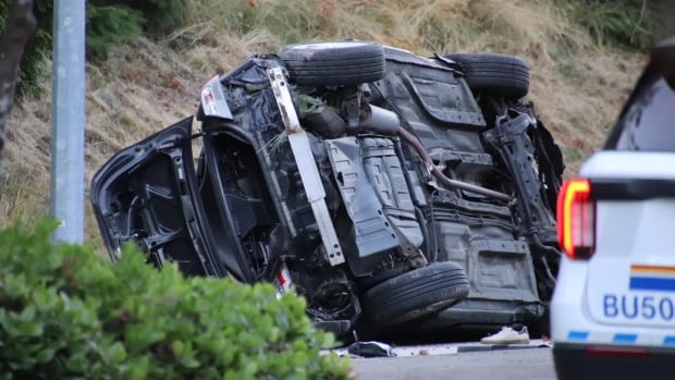 A vehicle that appears to be a black SUV is turned over on its side, its underside visible in the image. The corner of a white police vehicle is seen on the bottom right-hand corner of the image.