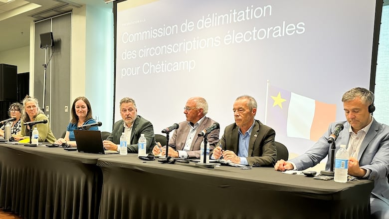Seven men and women are seated at a long table with microphones and a large screen behind them.