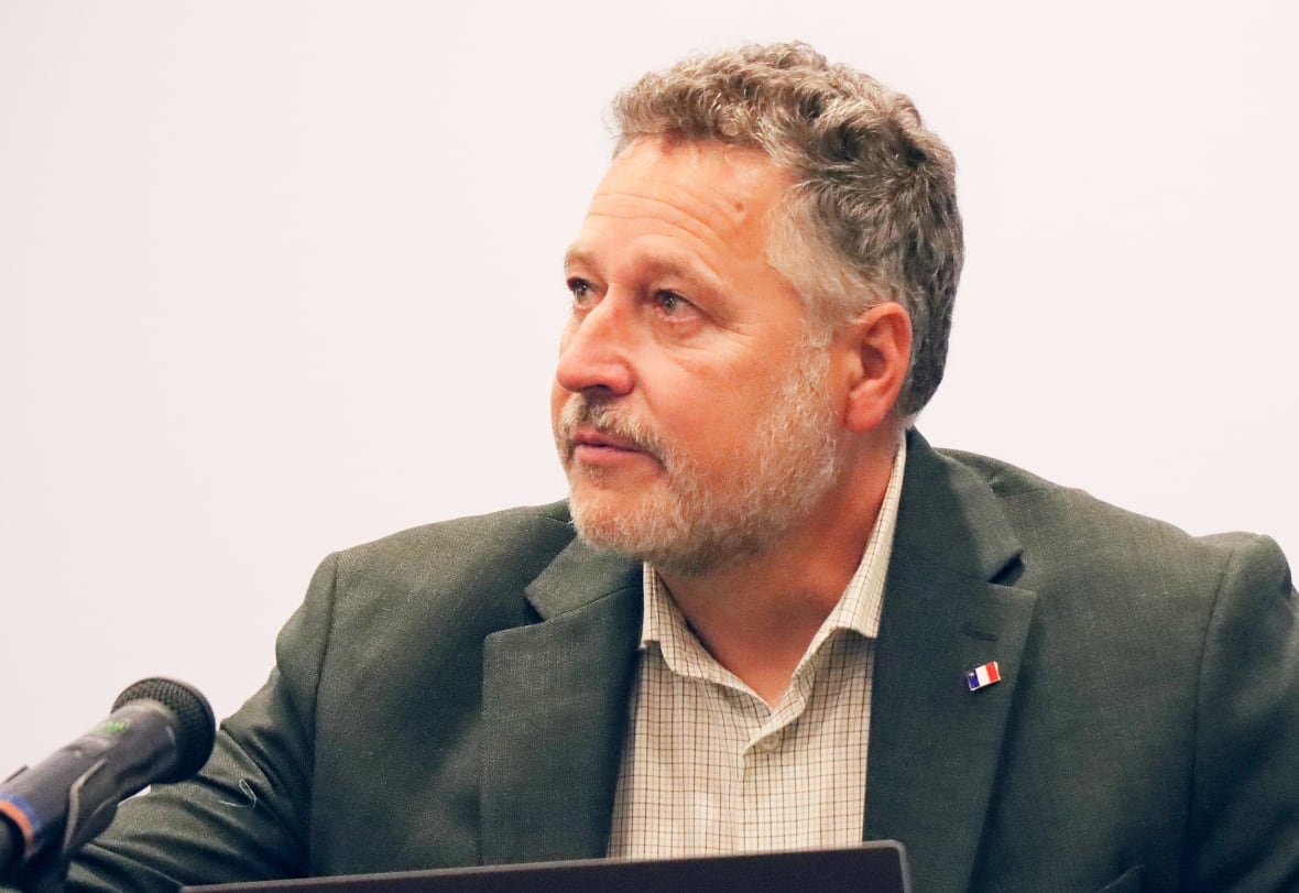 A man with curly grey hair and a grey beard and moustache wearing a dark green suit jacket and open-necked shirt sits in front of a microphone.