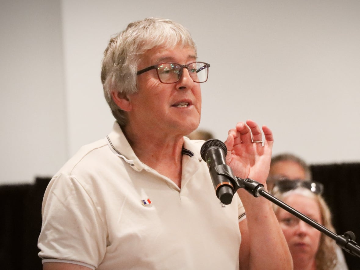 A man with grey hair and glasses wearing an off-white polo shirt gestures with his hand while speaking into a microphone.