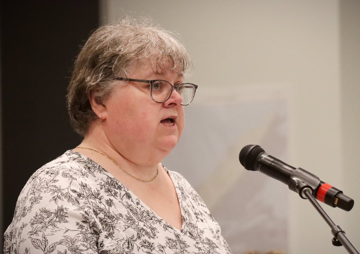 A woman with grey hair wearing a white flower-patterned blouse speaks into a microphone.