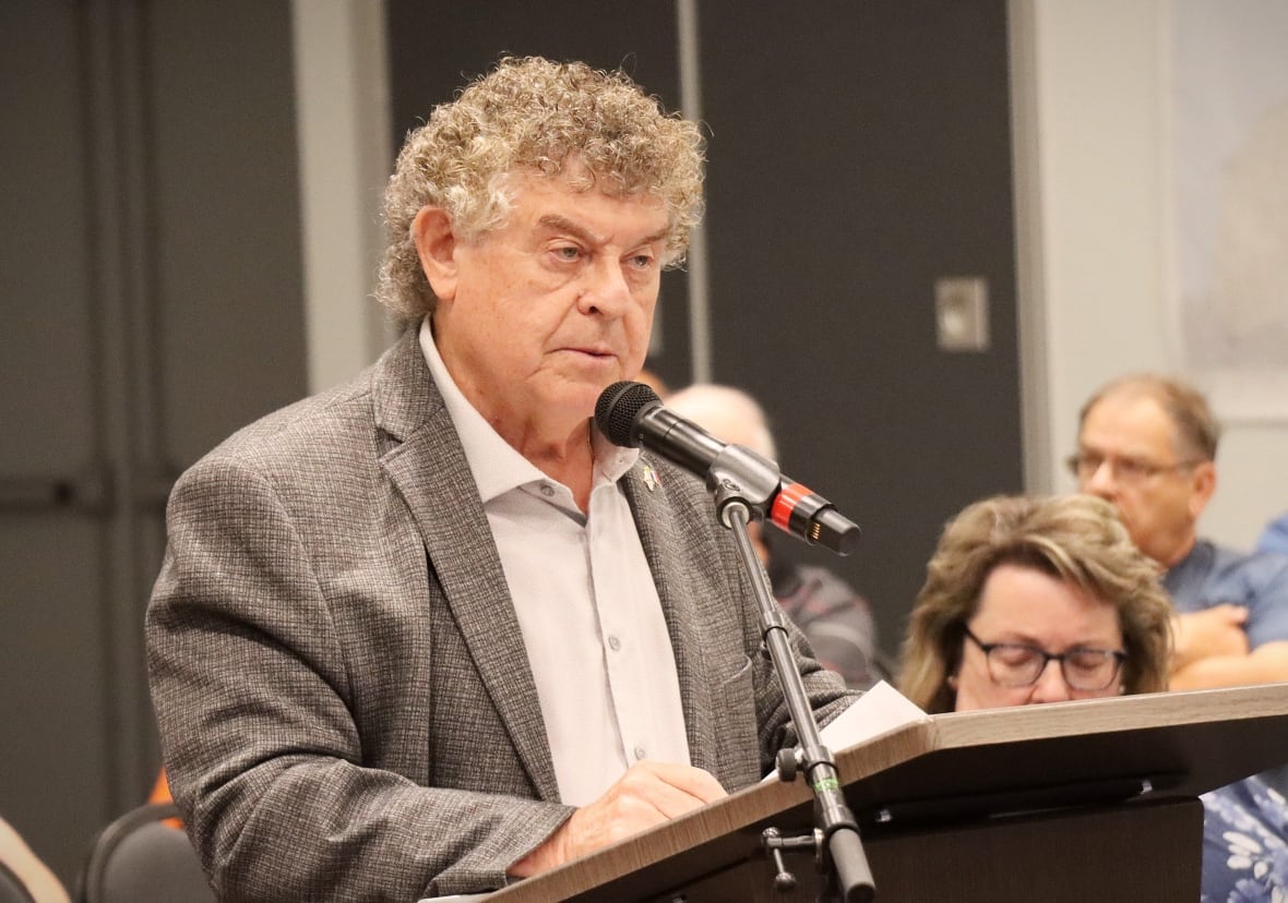 A man with curly grey hair wearing a grey suit and off-white open-necked shirt speaks into a microphone.