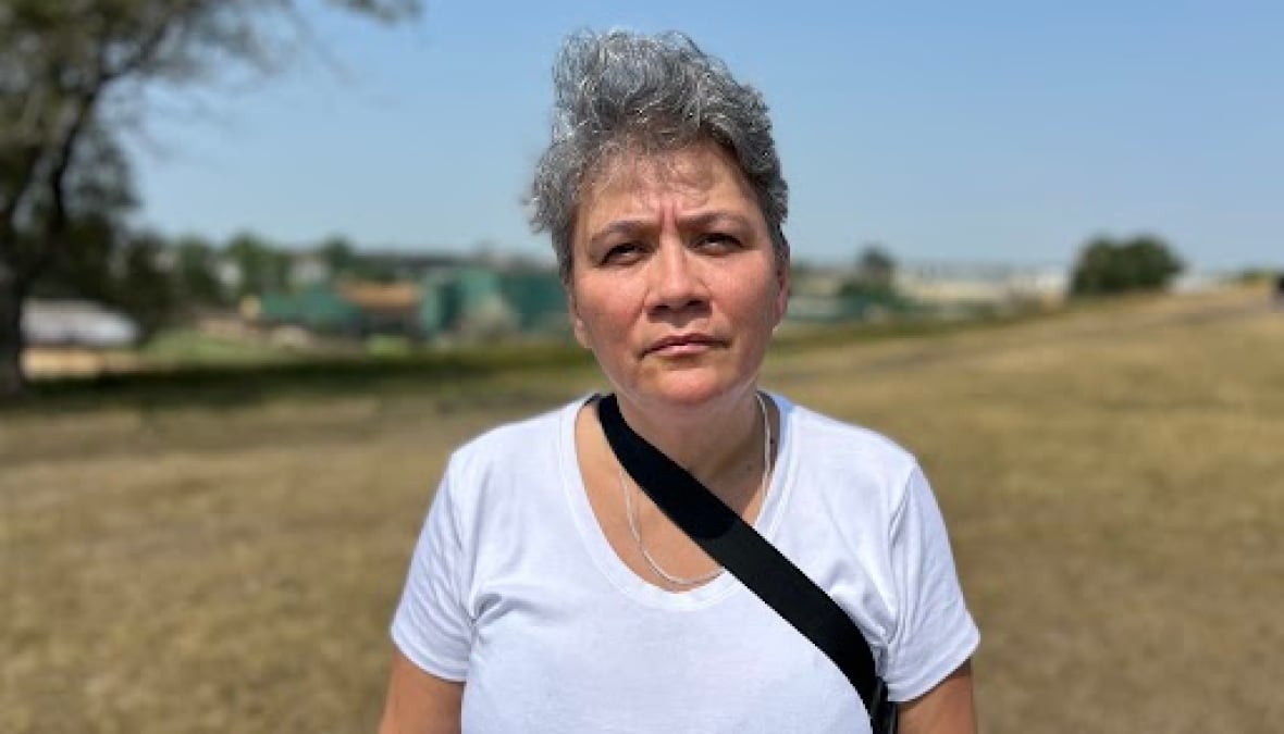 A woman with grey hair stands outside. She wears a white T-shirt.