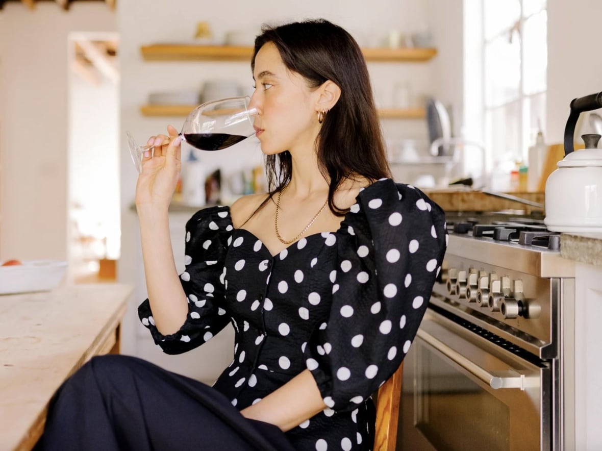 A woman sitting in a kitchen drinking a glass of wine. She's wearing a black blouse with white polka dots.