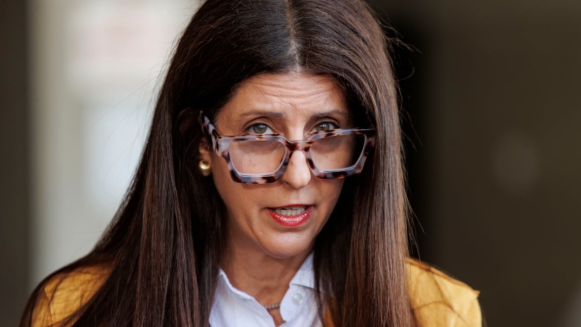 A woman with long brown hair, large glasses and a yellow blazer speaks into a microphone.