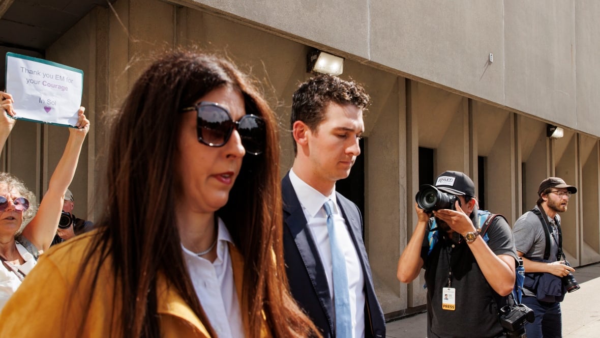 A man in a blue suit and tie walks with his lawyer, who is wearing sunglasses and a yellow blazer.