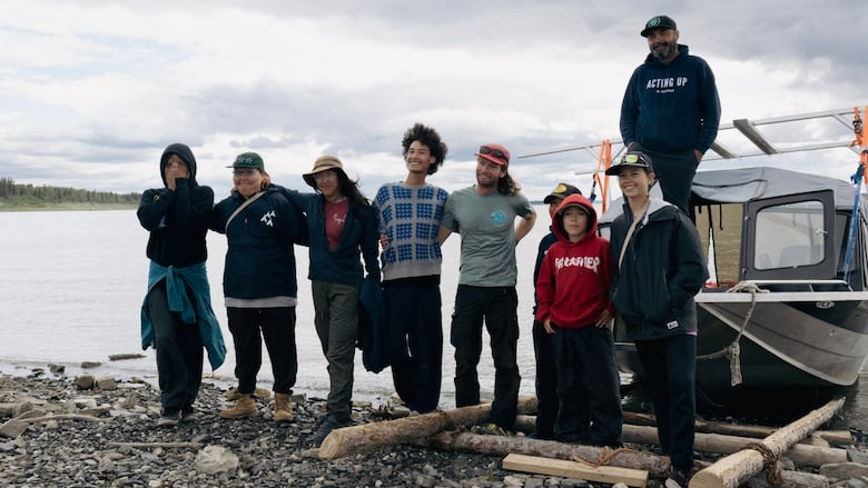Nine people stand smiling in front of a boat on the shore of an expanse of water.