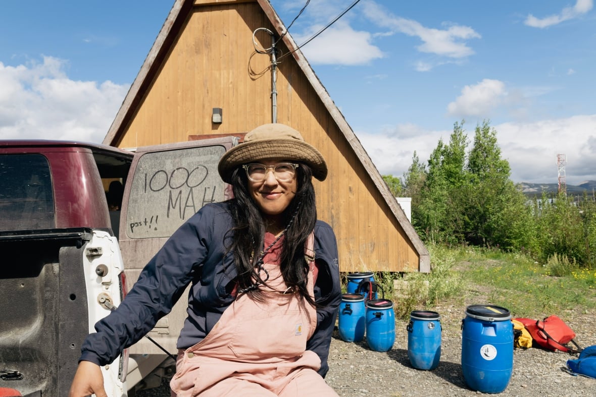 A young women in pink overalls and a tan hat sits in front of a dusty vehicle that has words written in the dirt on its window.