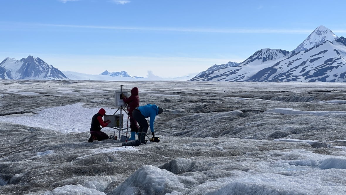 Three people standing on an icefield with snowcapped mountains in the background. 