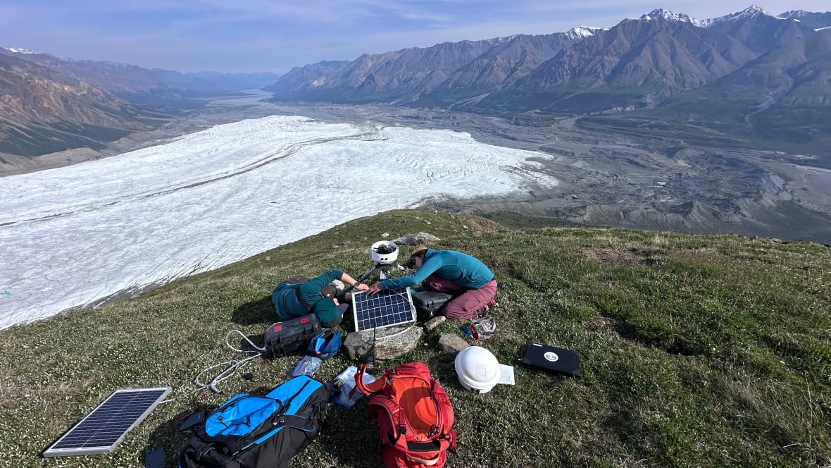 Two people on a patch of grass setting up technical equipment with an glacier and mountains in the background. 