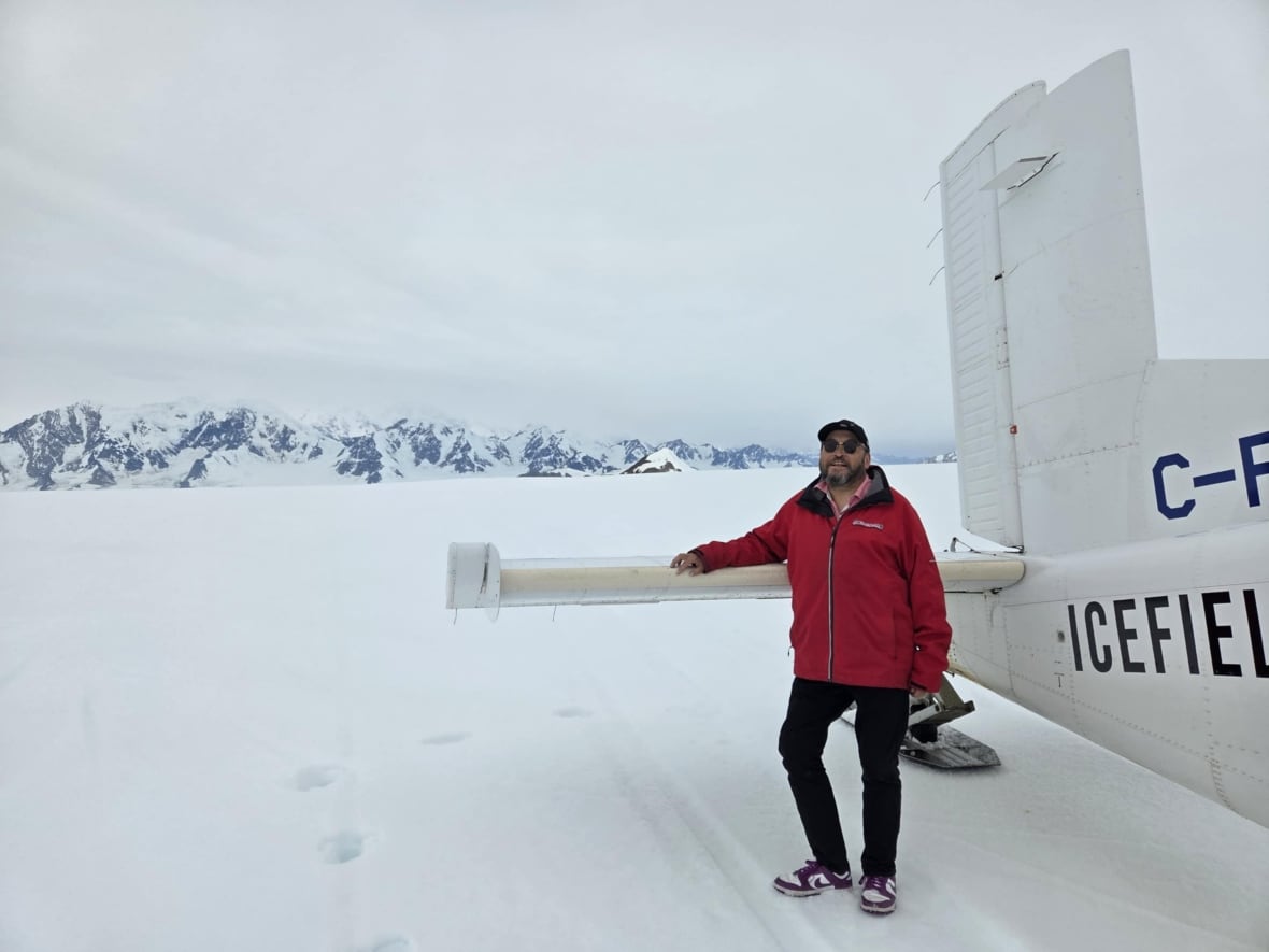 A man standing next to a plane on snow with a large snowcapped mountain in the background.