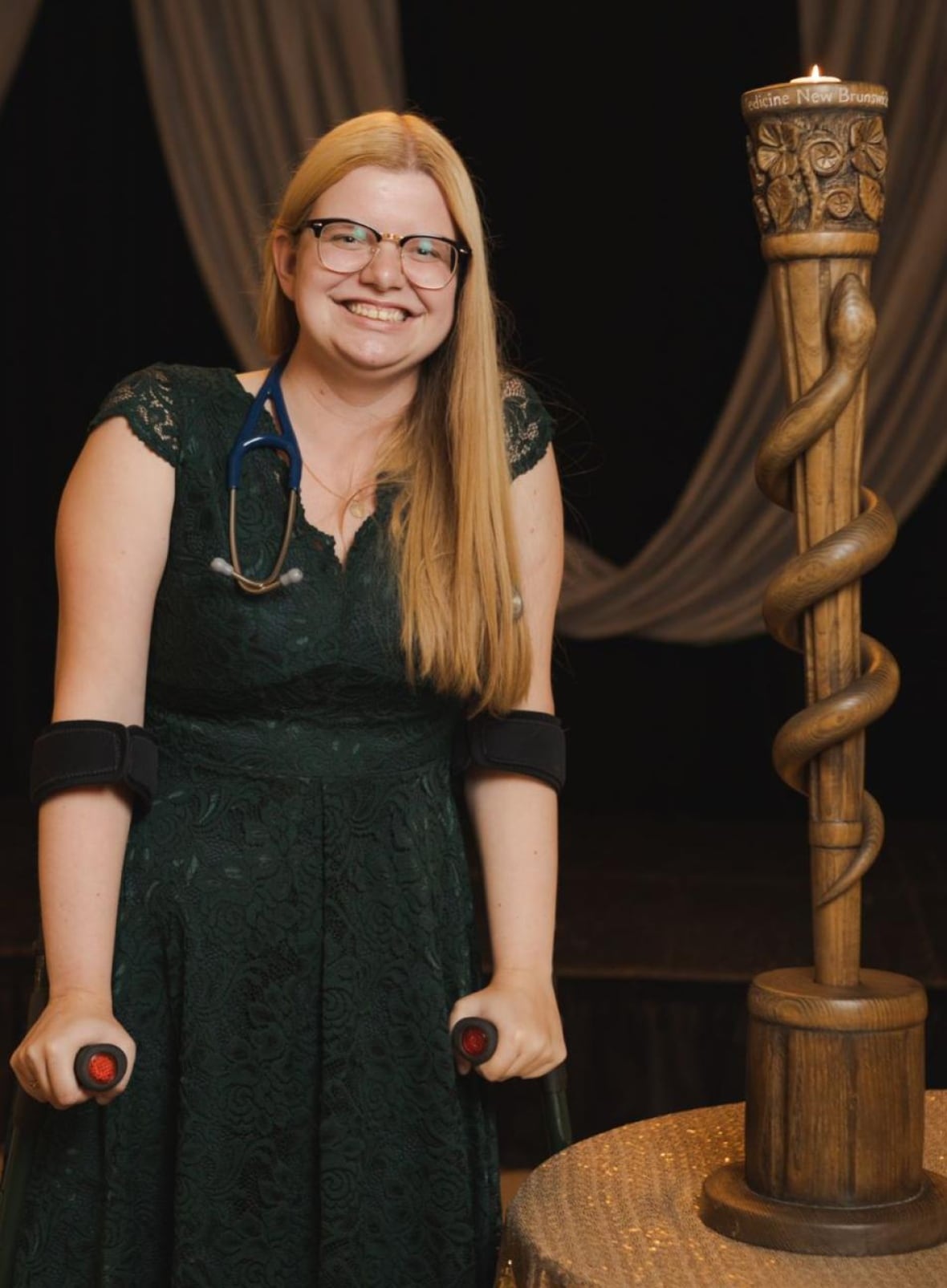 A smiling woman with blond hair poses for a photo. She’s wearing a green lace dress and a stethoscope around her neck. She’s using crutches and standing next to a wooden staff with a snake around it.